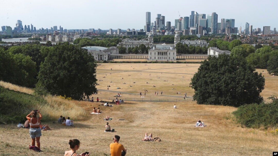Gente sentada en pasto seco por el sol en el Parque de Greenwich, con el Museo Marítimo y el distrito financiero de Canary Wharf al fondo, en Londres, el domingo 17 de julio de 2022. 