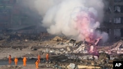 Bomberos en trajes protectores observan humo parcialmente rosado saliendo de las explosiones en un depósito en el noreste de China.