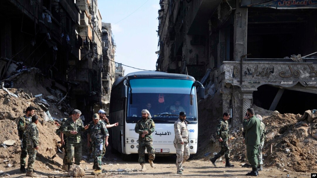 FILE - In this photo released on April 30, 2018 by the Syrian official news agency SANA, Syrian government forces gather in front of a bus carrying al-Qaida-linked fighters during an evacuation from the Palestinian refugee camp 0f Yarmuk, near Damascus. 