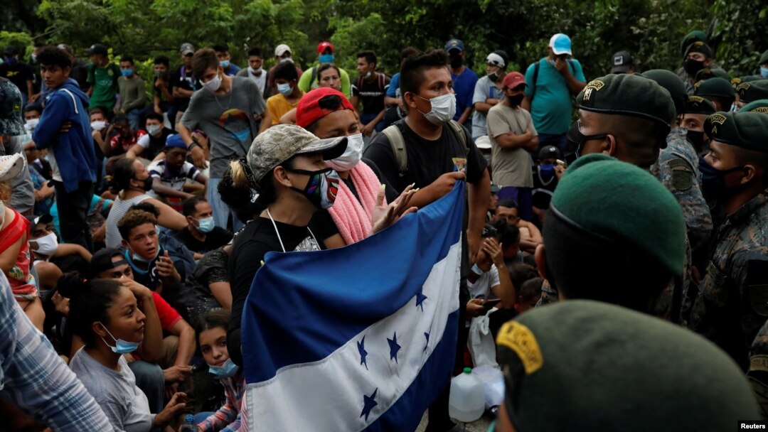 Migrantes de Honduras sostienen una bandera hondureña mientras están parados frente a soldados guatemaltecos que bloquean una carretera para evitar que los migrantes lleguen a la frontera de México, en San Pedro Cadenas, Izabal, Guatemala, octubre 2020.